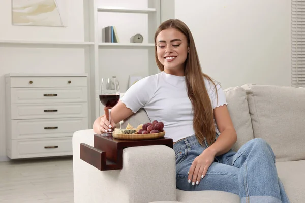 Happy woman holding glass of wine at home. Snacks on sofa armrest wooden table