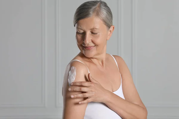 Happy woman applying body cream on shoulder near white wall