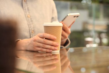 Masada elinde kağıt bardak ve akıllı telefon tutan bir kadın var. Yakın plan. Kahve hazır.