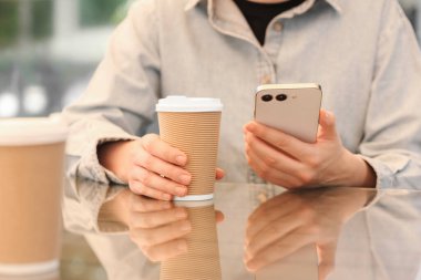 Masada elinde kağıt bardak ve akıllı telefon tutan bir kadın var. Yakın plan. Kahve hazır.