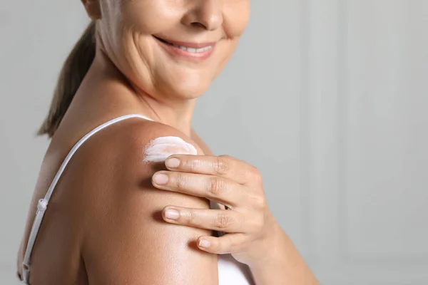 Happy woman applying body cream onto shoulder near white wall, closeup. Space for text