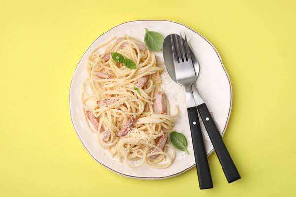 Plate of tasty pasta Carbonara with basil leaves, fork and spoon on yellow background, top view