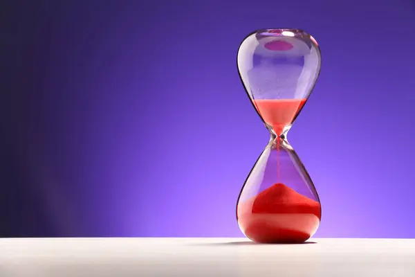 Hourglass with red flowing sand on white table against purple background. Space for text