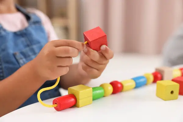 Motor skills development. Little girl playing with wooden pieces and string for threading activity at table indoors, closeup