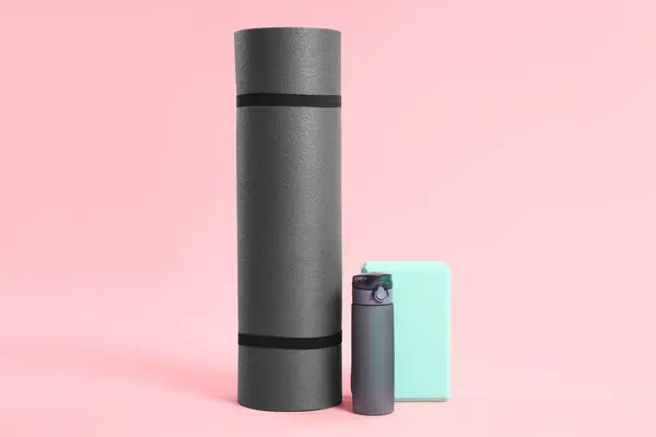 Grey exercise mat, yoga block and bottle of water on pink background