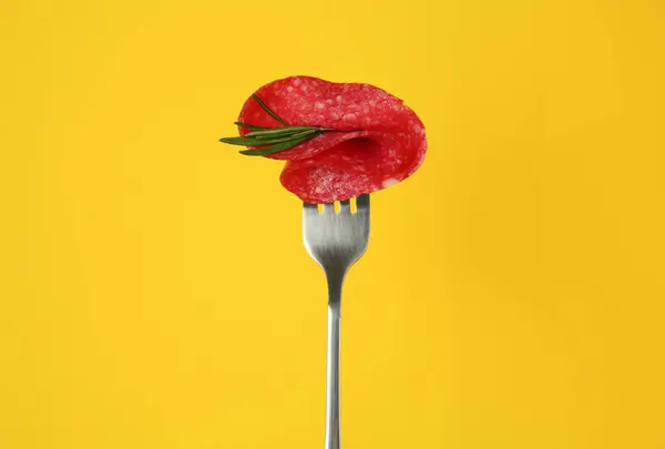 Fork with tasty slice of salami and rosemary on orange background