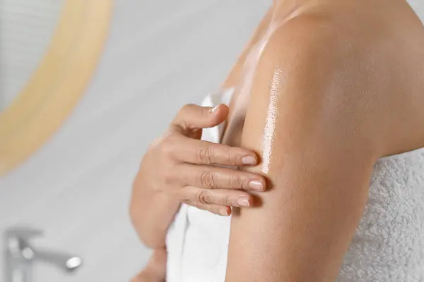 Woman applying body oil onto shoulder in bathroom, closeup. Space for text