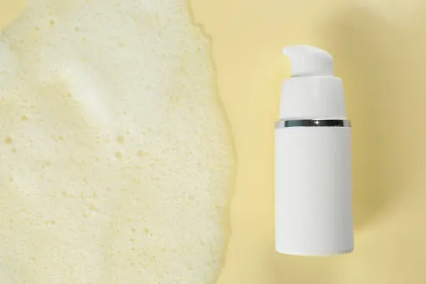Bottle of face cleanser and white foam on beige background, top view with space for text. Skin care cosmetic