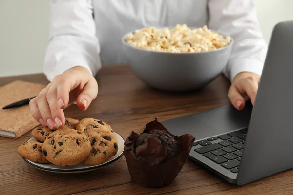 Bad habits. Woman eating cookies while using laptop at wooden table, closeup