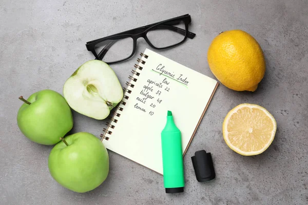 Notebook with products of low glycemic index, marker, glasses and fresh fruits on grey table, flat lay