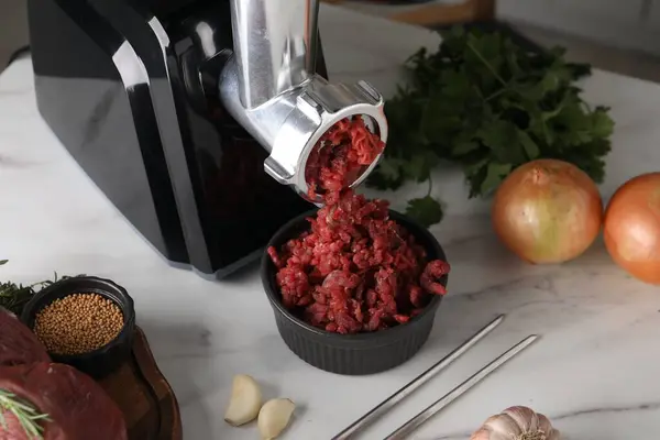 Electric meat grinder with beef mince and products on white marble table in kitchen