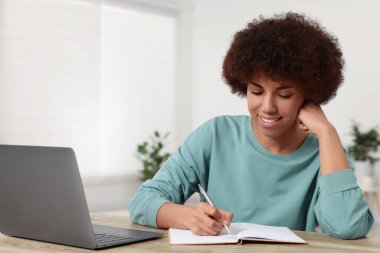 Genç bir kadın, odasındaki tahta masada dizüstü bilgisayar kullanıyor ve deftere yazıyor.