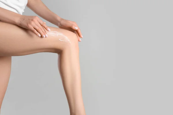 Woman applying body cream onto her smooth legs on light grey background, closeup. Space for text