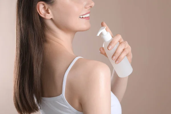 Woman applying body oil onto shoulder on beige background, closeup