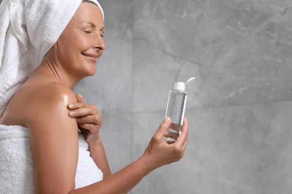 Happy woman applying body oil onto shoulder near grey wall