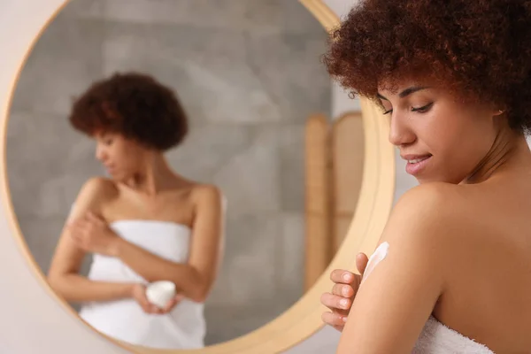 Beautiful young woman applying body cream onto arm indoors