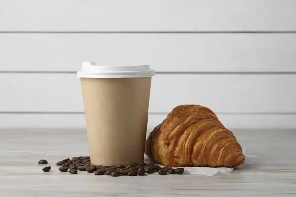 Coffee to go. Paper cup with tasty drink, croissant and beans on white wooden table