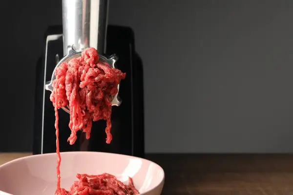 Electric meat grinder with beef mince on wooden table against grey background, closeup. Space for text