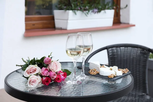 Bouquet of roses, glasses with wine and food on glass table near house on outdoor terrace