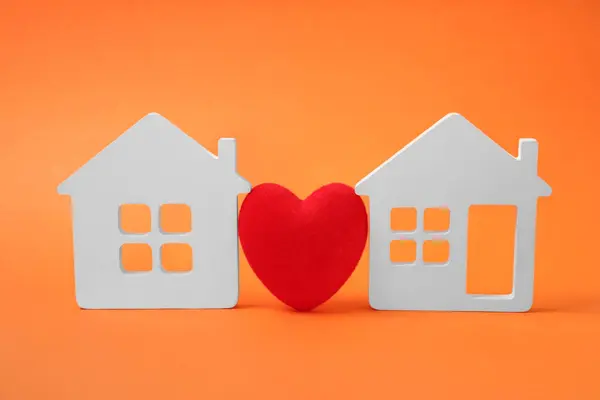 Long-distance relationship concept. Decorative heart between two white house models on orange background