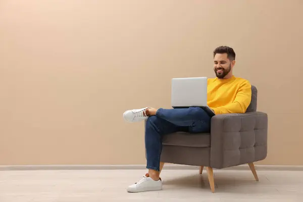 Handsome man with laptop sitting in armchair near beige wall indoors, space for text