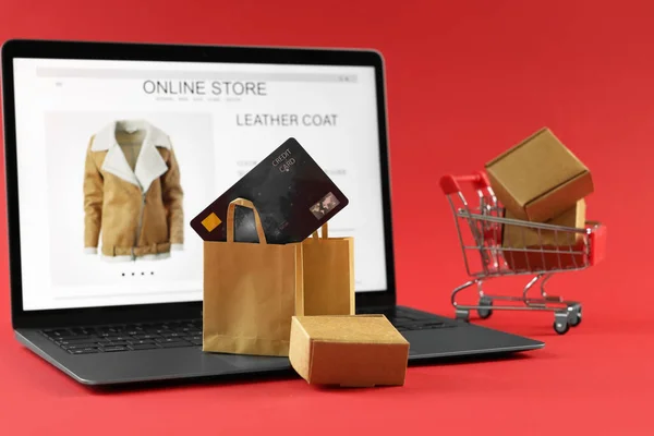 Online store. Laptop, small purchases, credit card and mini shopping cart on red background, selective focus
