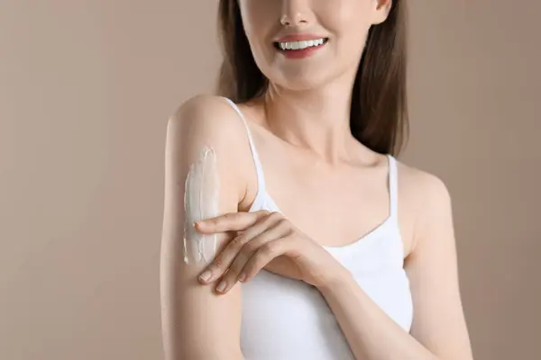 Woman applying body cream onto arm on beige background, closeup