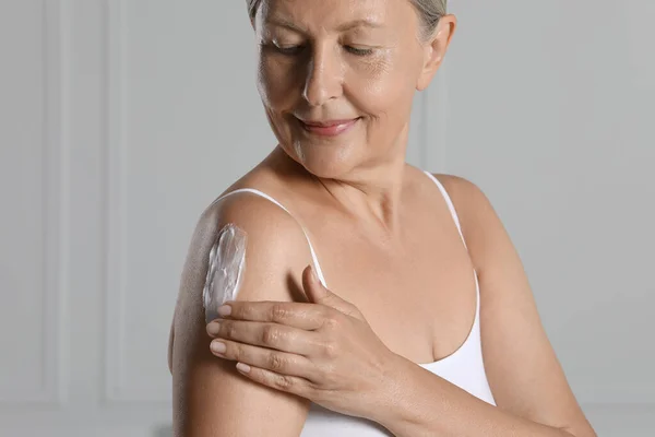 Happy woman applying body cream on shoulder near white wall