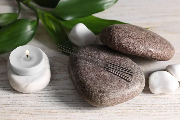 Stones with acupuncture needles and burning candle on white wooden table