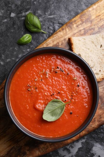 Delicious tomato cream soup in bowl and basil leaves on dark textured table, flat lay