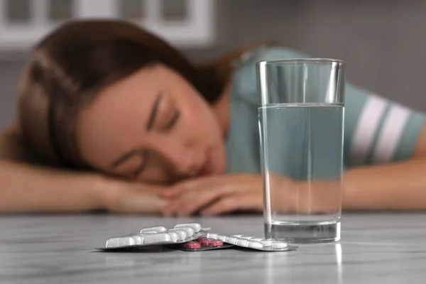 Woman sleeping at table with antidepressant pills and glass of water indoors, selective focus
