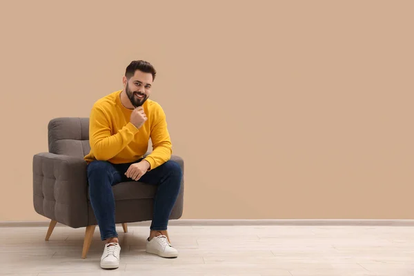 Handsome man sitting in armchair near beige wall indoors, space for text