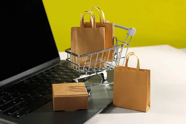 Online store. Laptop, mini shopping cart and purchases on beige table, closeup