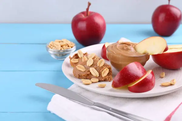 Slices of fresh apple with peanut butter, nuts and knife on light blue wooden table