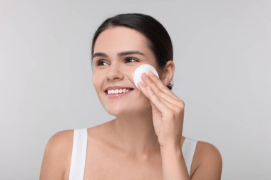 Genç bir kadın açık gri arka planda pamukla yüzünü temizliyor.