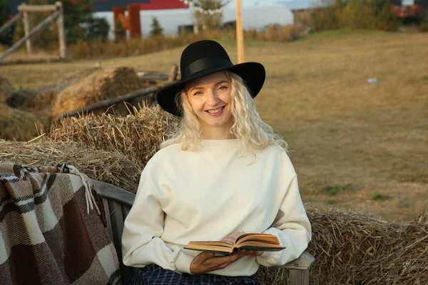 Beautiful woman with book sitting on wooden bench near hay bales outdoors. Autumn season