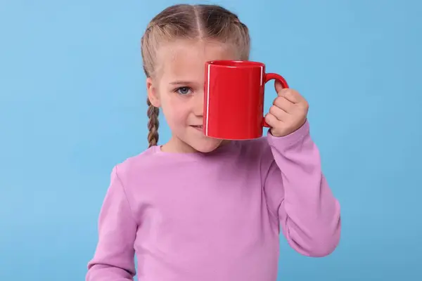 Cute girl covering eye with red ceramic mug on light blue background