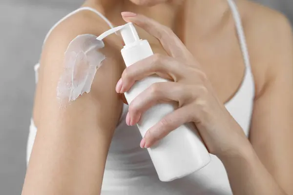 Woman applying body cream onto shoulder on grey background, closeup