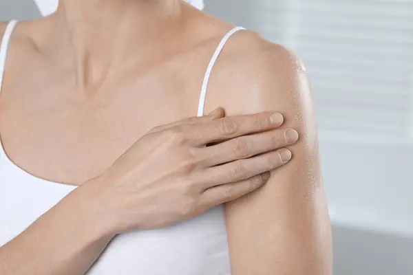 Woman applying body oil onto arm indoors, closeup