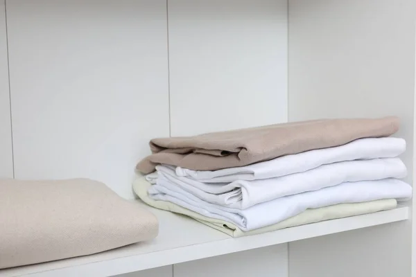 Stack of different folded shirts on white shelf. Organizing clothes