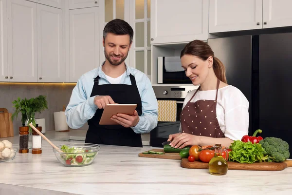 Happy couple reading recipe on tablet while cooking in kitchen. Online culinary book