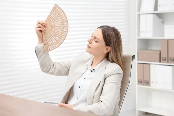 Businesswoman waving hand fan to cool herself at table in office