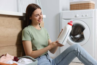 Çamaşır makinesinin yanında oturan ve banyoda yumuşatıcı tutan bir kadın.
