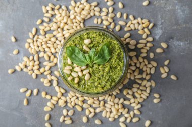 Lezzetli pesto soslu kavanoz, çam fıstığı ve fesleğen yaprağı gri masada, düz yerde.
