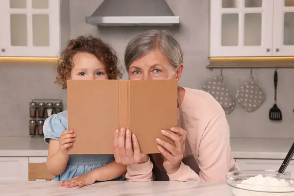 Cute little girl and her granny with recipe book in kitchen