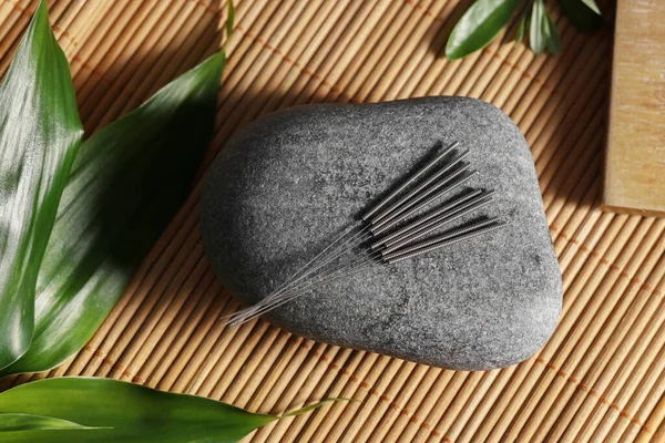 Stone with acupuncture needles and green leaves on bamboo mat, flat lay