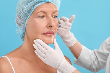 Doktor açık mavi arka planda yaşlı kadına dudak iğnesi yapıyor. Estetik ameliyat