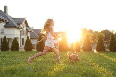 Güzel kız arka bahçede gün batımında yeşil çimlerde sevimli bir Maltipoo köpeğiyle koşuyor.