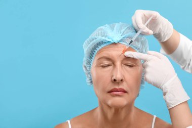 Doktor, açık mavi arka planda yaşlı kadına yüz enjeksiyonu yapıyor. Mesaj için yer var. Estetik ameliyat