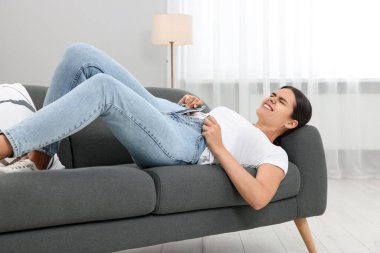 Evdeki kanepede yatarken dar kot pantolon giymek için çabalayan genç bir kadın.